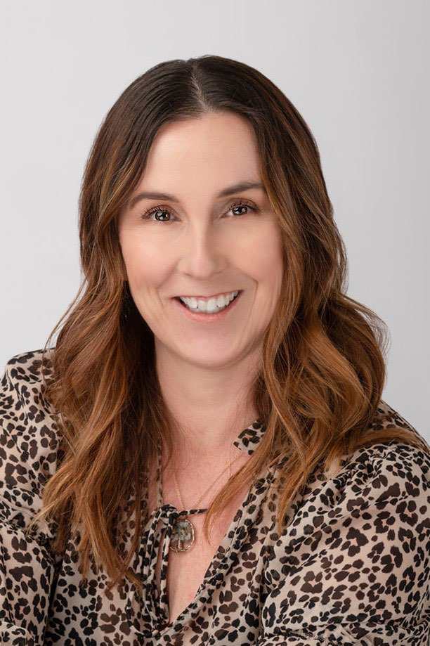A smiling woman with shoulder-length brown hair wearing a beige leopard-print blouse, photographed against a plain light gray background.